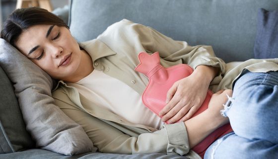 Junge Frau ruht sich auf einem Sofa aus, eine Wärmeflasche auf dem Bauch.