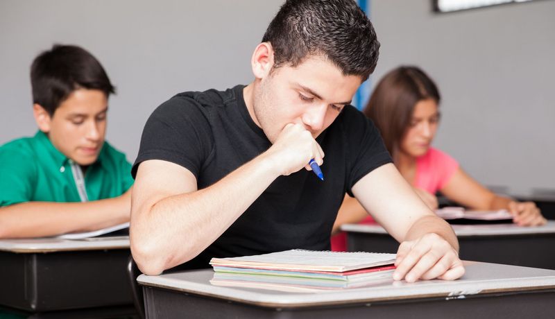 Ein Jugendlicher brütet in der Schulklasse über seinen Aufgaben, er hält den Stift in einer Faust nahe am Mund.. Im Hintergrund sind ein Mitschüler und eine Mitschülerin zu sehen.