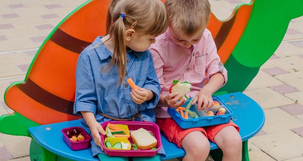 Zwei Kinder, sitzen auf einer Bank und essen Pausenbrot.