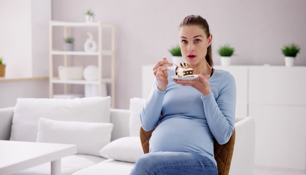 Eine schwangere Frau schaut erstaunt, in der Hand ein Tellerchen mit einem Stück Kuchen darauf.