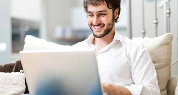 Junger lachender Mann mit dunklen Haaren und dunklem Bart auf der Couch sitzend, einen Laptop auf den Knien.