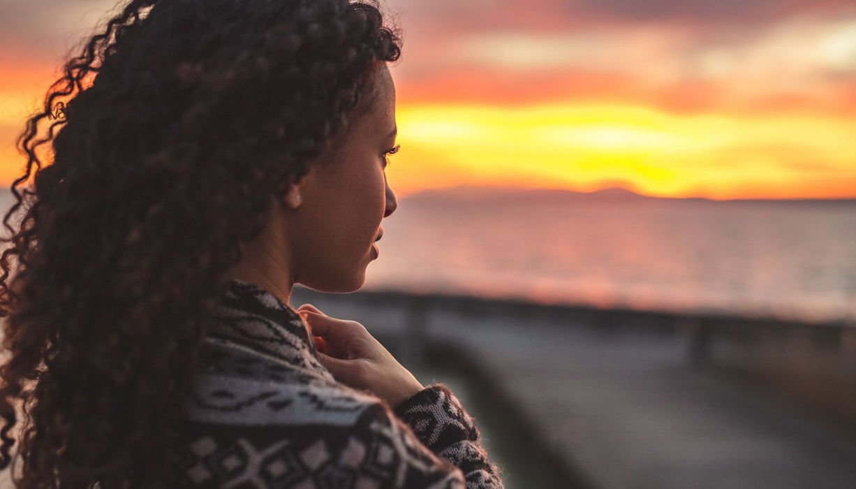 Junge Frau mit dunklen Locken, schaut in die Ferne. Im Hintergrund Sonnenuntergang und Meer. 