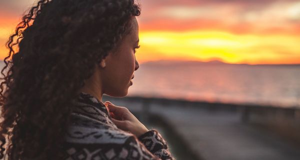 Junge Frau mit dunklen Locken, schaut in die Ferne. Im Hintergrund Sonnenuntergang und Meer. 