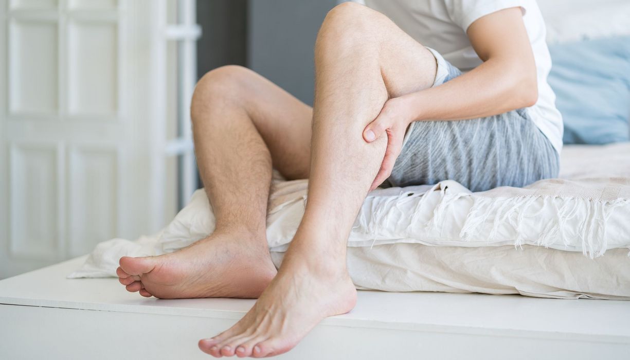 Junger Mann sitzt auf dem Bett und fasst sich an die Wade.