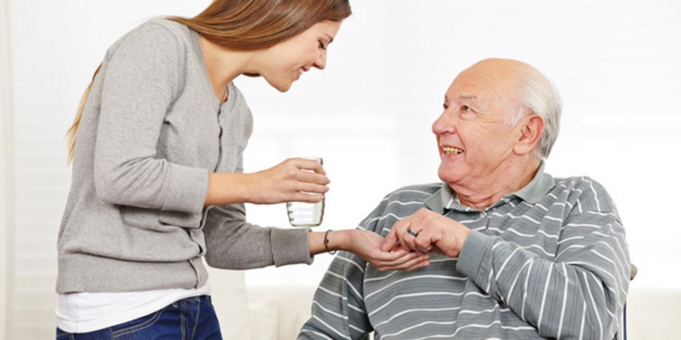Senior (Mann) im Rollstuhl, graugestreifter Pullover, schaut lächelnd zu jüngerer Frau (graue Feinstrickjacke, Jeans) auf, die ihm ein Glas Wasser und Tabletten reicht