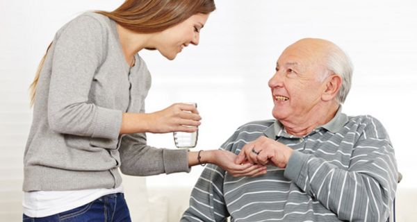 Senior (Mann) im Rollstuhl, graugestreifter Pullover, schaut lächelnd zu jüngerer Frau (graue Feinstrickjacke, Jeans) auf, die ihm ein Glas Wasser und Tabletten reicht