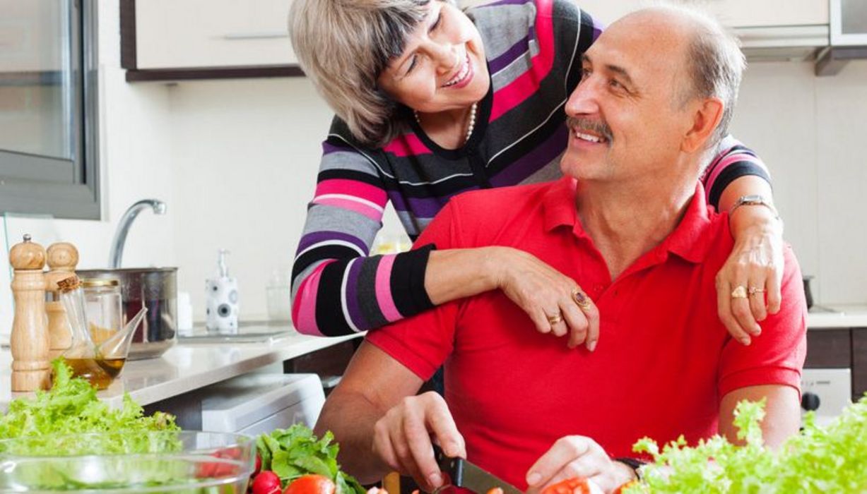 Gut gelaunte Senioren, ca. Ende 60, sportliche Freizeitklamotten, in der Küche beim Salatschneiden. Er sitzt und schaut zu ihr hoch. Sie umarmt ihn lachend von hinten