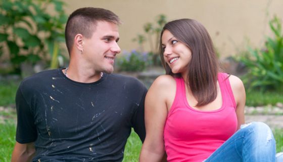 Junges Paar auf einer Wiese nebeneinander sitzend (Jeans, Shirts) schaut sich verliebt in die Augen