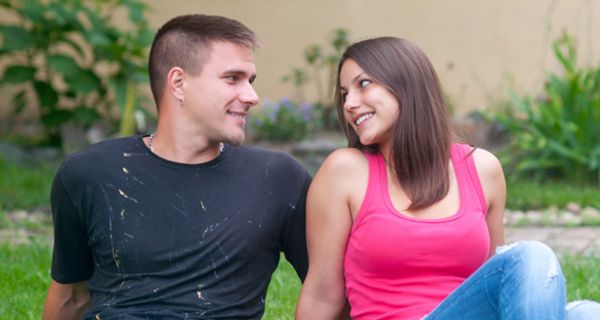 Junges Paar auf einer Wiese nebeneinander sitzend (Jeans, Shirts) schaut sich verliebt in die Augen