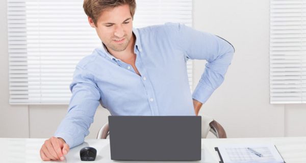 Junger Mann sitzt an Bürotisch vor einem Laptop und hält sich den schmerzenden Rücken