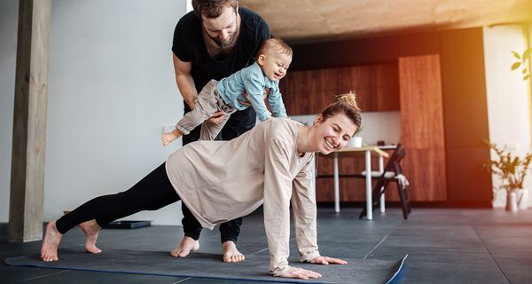 Frau macht auf dem Boden eine Plank, Mann setzt ihr ein kleines Kind auf den Rücken.