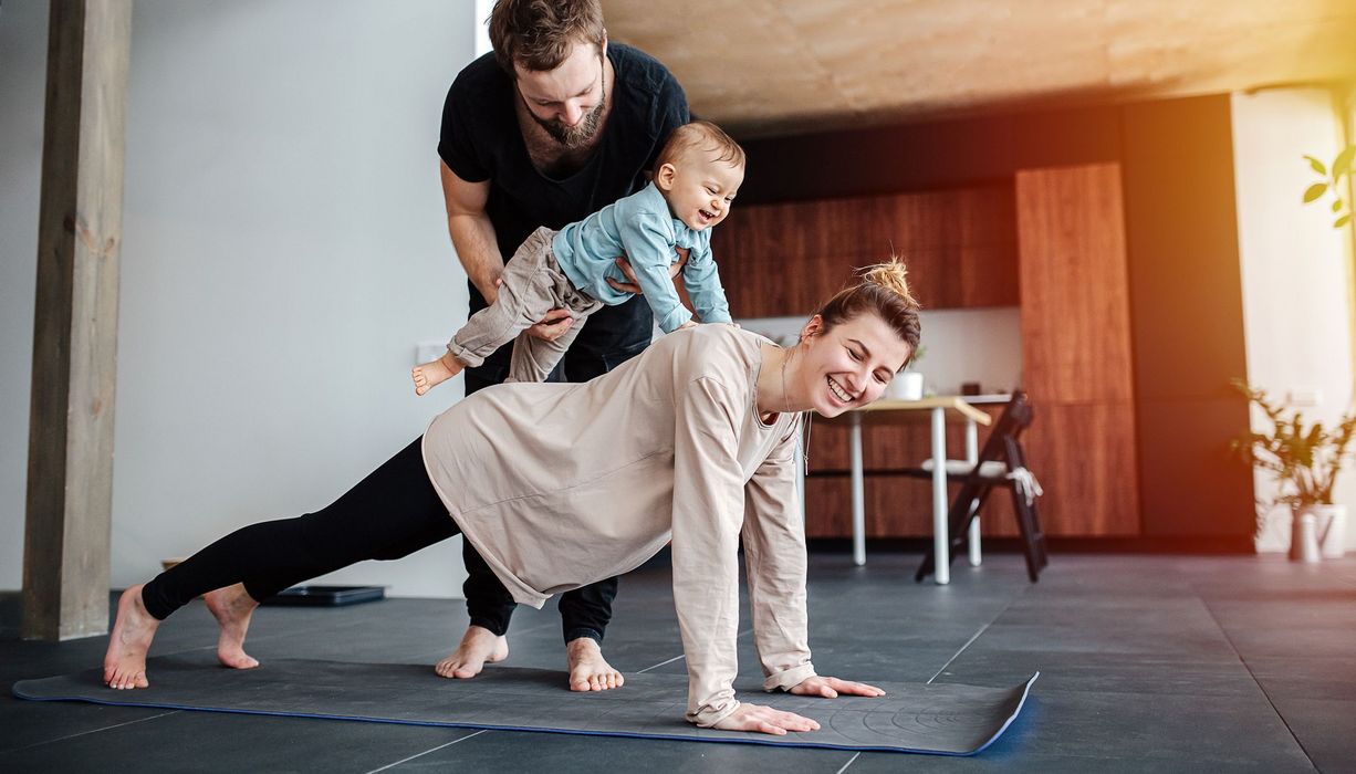 Frau macht auf dem Boden eine Plank, Mann setzt ihr ein kleines Kind auf den Rücken.