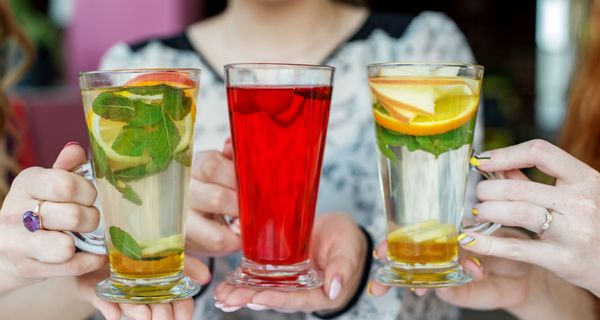 Bildzentrum: Zwei Minz-Orange-Tees und ein roter Früchtetee in den Händen von jungen Frauen (verschwommen im Hintergrund).