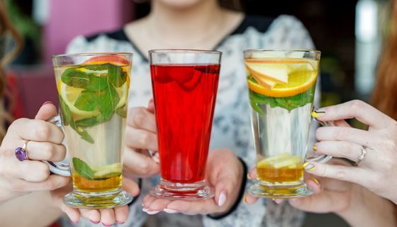Bildzentrum: Zwei Minz-Orange-Tees und ein roter Früchtetee in den Händen von jungen Frauen (verschwommen im Hintergrund).