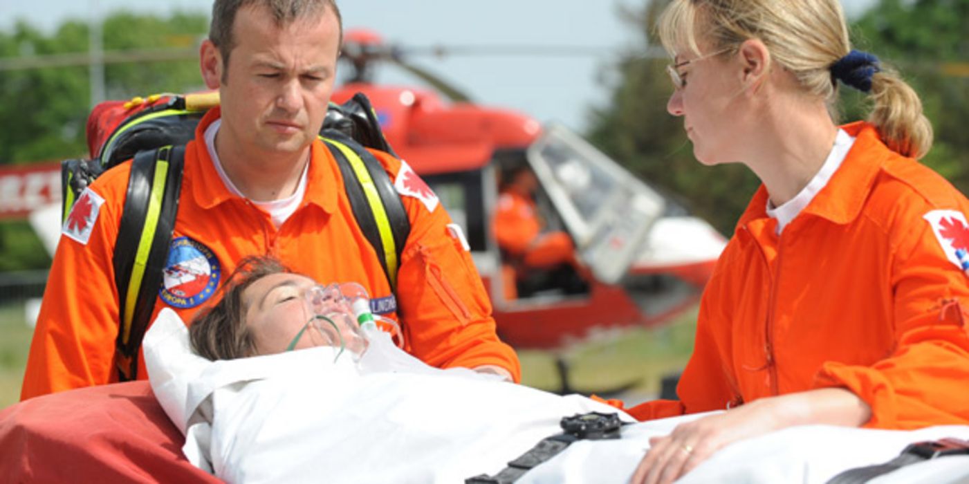 Szene mit Notfallhelferin und -helfer in oranger Kleidung, Frau auf Trage festgeschnallt mit Atemmaske, im Hintergrund Rettungshubschrauber