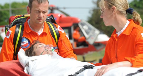 Szene mit Notfallhelferin und -helfer in oranger Kleidung, Frau auf Trage festgeschnallt mit Atemmaske, im Hintergrund Rettungshubschrauber