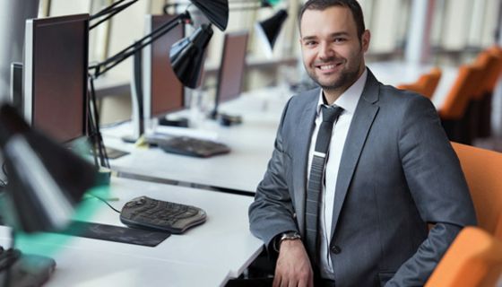 Jüngerer Mann in den 20ern, taubenblauer Anzug, weißes Hemd, gemusterte Krawatte, kurze, dunkle Haare und Bart, linker Unterarm auf Schreibtisch, lächelt in die Kamera