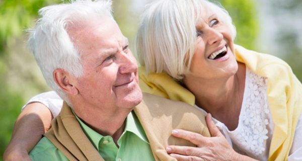 Zwei Senioren, Frau hält Mann im Arm, strecken ihr Gesicht in die Sonne und lachen.