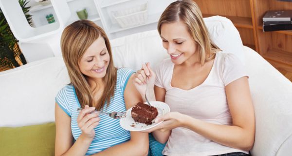 Zwei vergnügte Frauen sitzen auf einer Couch im Wohnzimmer und naschen beide an einem Stück Torte, das die eine auf einem Teller in der Hand hält