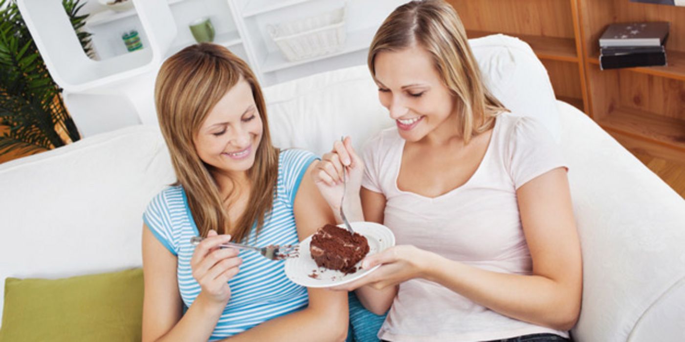 Zwei vergnügte Frauen sitzen auf einer Couch im Wohnzimmer und naschen beide an einem Stück Torte, das die eine auf einem Teller in der Hand hält