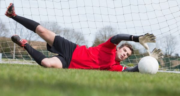 Torwart, rotes Shirt, ansonsten schwarze Kleidung, mit vollem Körpereinsatz, liegt im Tor, Ball vor seinen ausgestreckten Händen