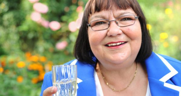 Frau, übergewichtig, ca. 50, Ponyfrisur halblang, Brille, Portrait, sommerblaue Jacke mit weißer Paspelierung, ein Glas mit Wasser in der rechten Hand, lacht freundlich in die Kamera