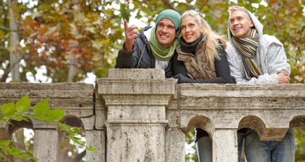 Junge Leute, 2 Männer, 1 Frau in warmer Kleidung stehen im Stadtpark an einer Steinbrüstung; im Hintergrund Bäume mit braungrünen Blättern