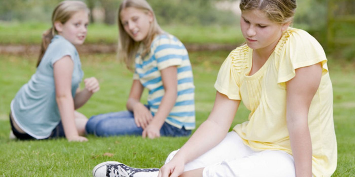 Etwas übergewichtiges Mädchen, ca. 10 Jahre alt, im hellgelben Kleid auf einer Wiese, im Hintergrund zwei gleichaltrige Mädchen in Jeans und T-Shirt, die miteinander flüstern