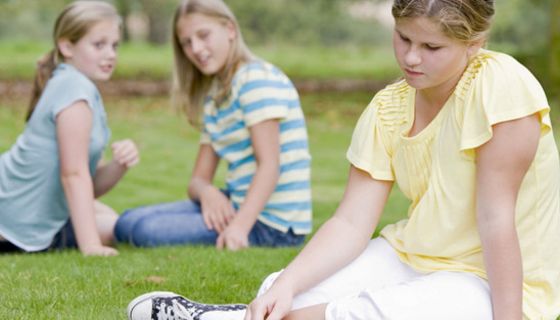 Etwas übergewichtiges Mädchen, ca. 10 Jahre alt, im hellgelben Kleid auf einer Wiese, im Hintergrund zwei gleichaltrige Mädchen in Jeans und T-Shirt, die miteinander flüstern