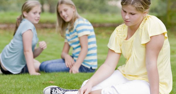 Etwas übergewichtiges Mädchen, ca. 10 Jahre alt, im hellgelben Kleid auf einer Wiese, im Hintergrund zwei gleichaltrige Mädchen in Jeans und T-Shirt, die miteinander flüstern