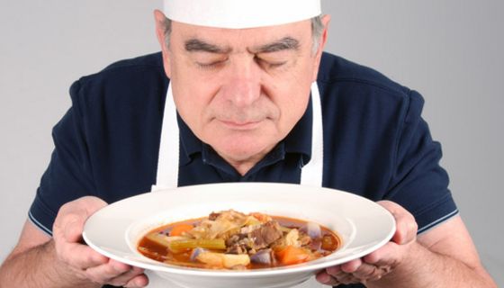 Mann in den 60ern, Kochmütze und weiße Schürze, hält in beiden Händen einen Suppenteller mit Rindfleisch und Brühe und schaut genießerisch hinein