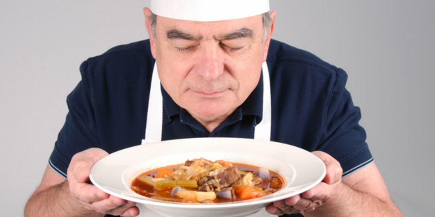 Mann in den 60ern, Kochmütze und weiße Schürze, hält in beiden Händen einen Suppenteller mit Rindfleisch und Brühe und schaut genießerisch hinein