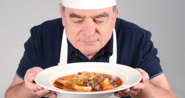 Mann in den 60ern, Kochmütze und weiße Schürze, hält in beiden Händen einen Suppenteller mit Rindfleisch und Brühe und schaut genießerisch hinein