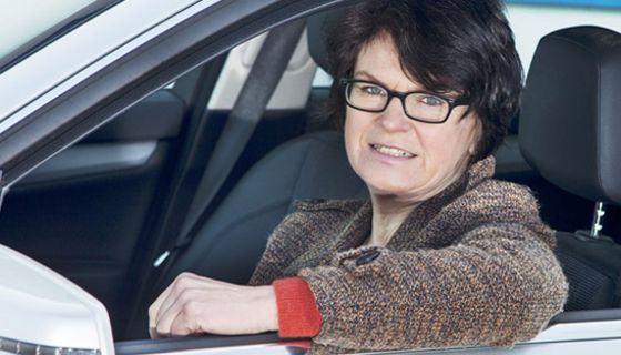 Frau um die 50, Brille, kurze, dunkle Haare, Hornbrille, am Steuer, linker Arm auf offenem Seitenfenster, in die Kamera blickend