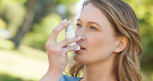 Nahaufnahme Gesicht, Schultern: Frau ca. Anfang 30, blonde Haare, blaue Bluse in Natur beim Benutzen eines Asthma-Sprays