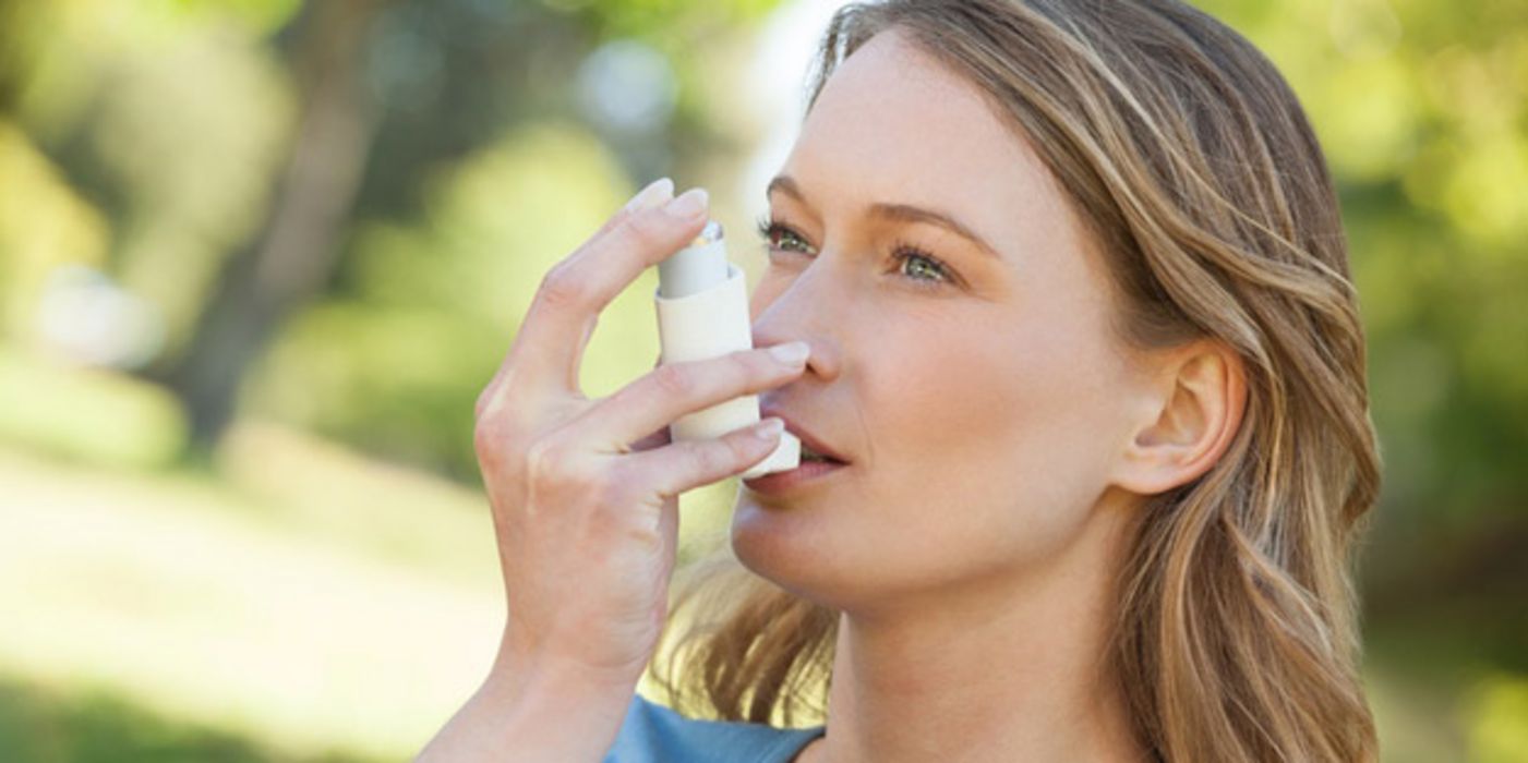 Nahaufnahme Gesicht, Schultern: Frau ca. Anfang 30, blonde Haare, blaue Bluse in Natur beim Benutzen eines Asthma-Sprays