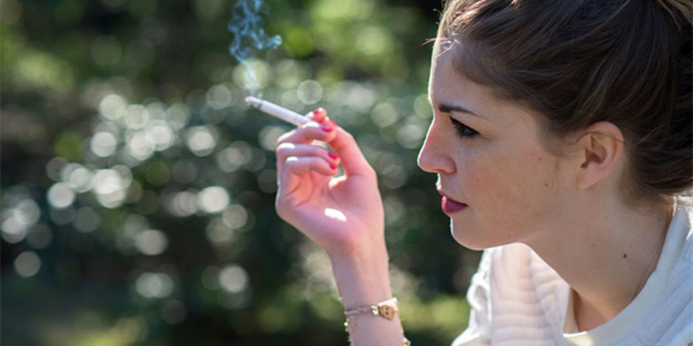 Rechter Bildrand, junge Frau im Halbprofil, Zigarette in rechter Hand, weißer Pulli, zusammengebundene Haare, Hintergrund Natur