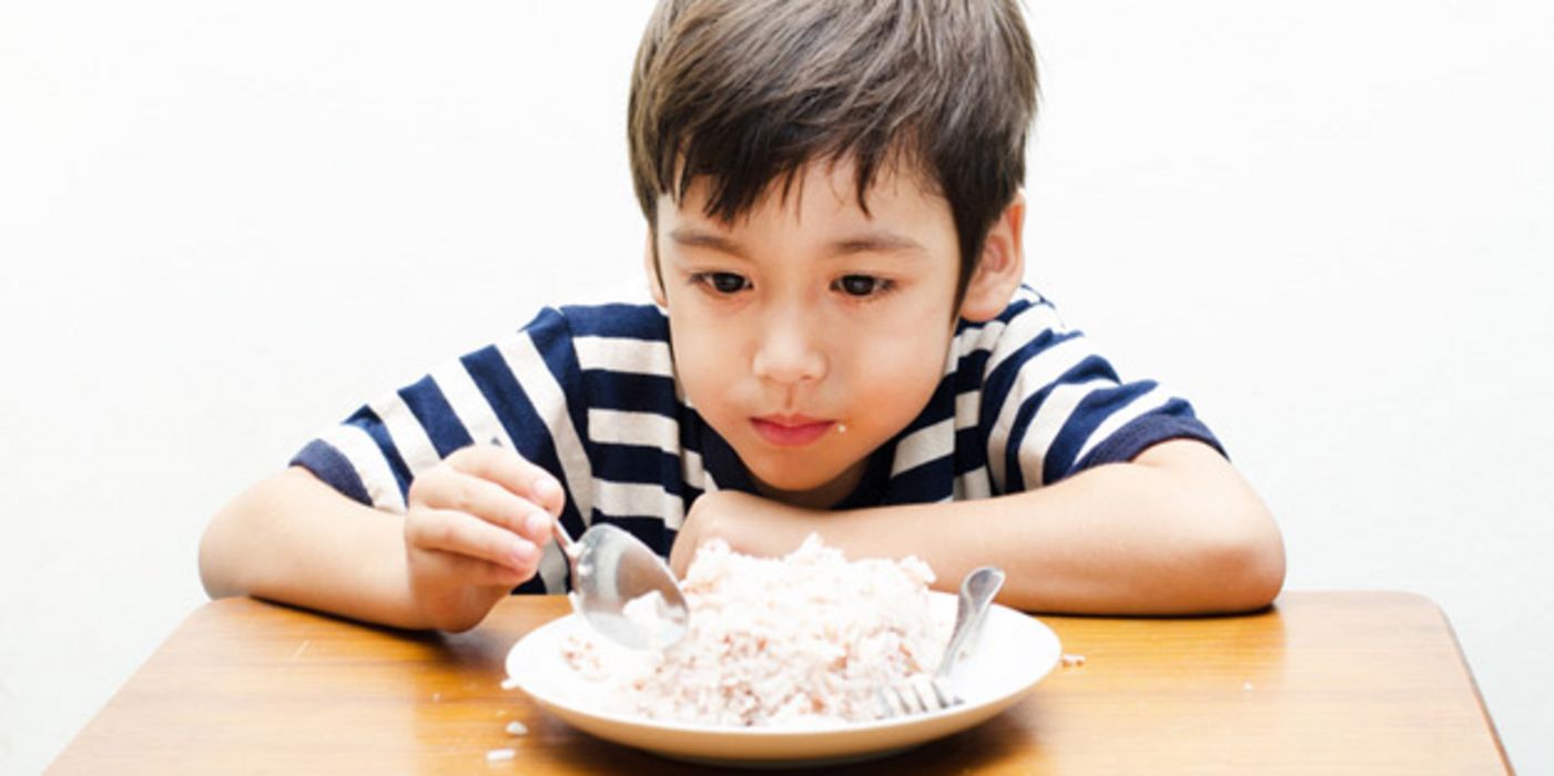 Kleiner Junge sitzt vor einem Teller mit Reis mit einem Löffel in der Hand.