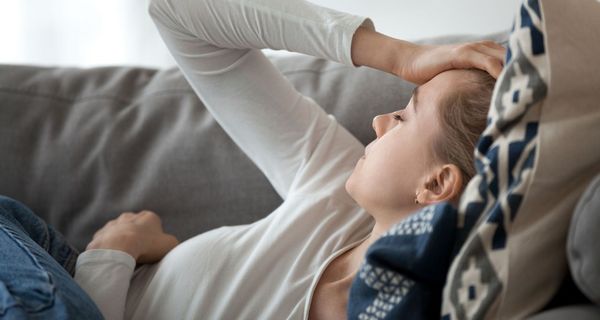 Junge Frau liegt auf der Couch und hält sich den Kopf, ist erschöpft.