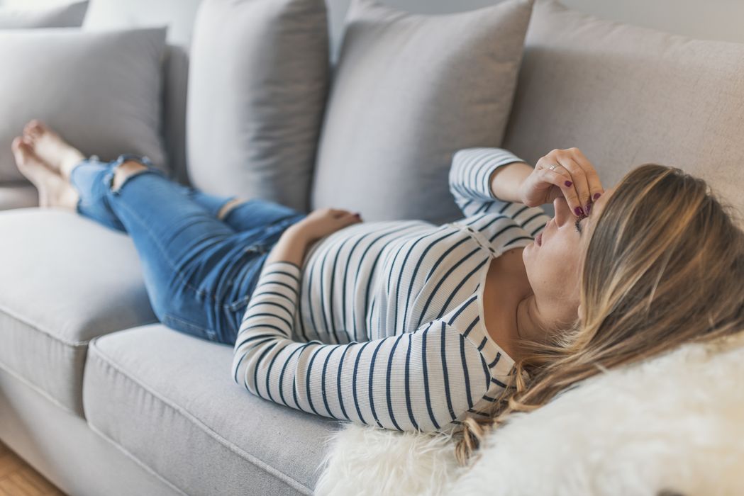 Junge Frau, liegt auf der Couch und hält eine Hand an ihre Schläfe.