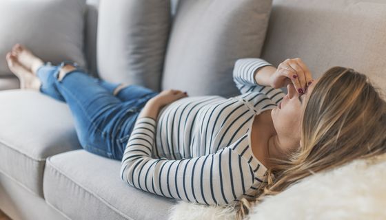 Junge Frau, liegt auf der Couch und hält eine Hand an ihre Schläfe.