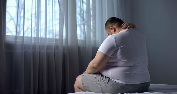 Fettleibiger Mann, sitzt mit dem Rücken zur Kamera auf einem Bett.