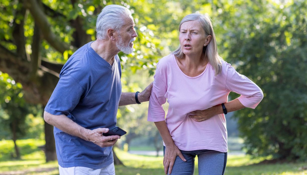 Ältere Frau hält sich den Oberbauch, ein Mann steht erschrocken neben ihr, das Handy in der Hand.