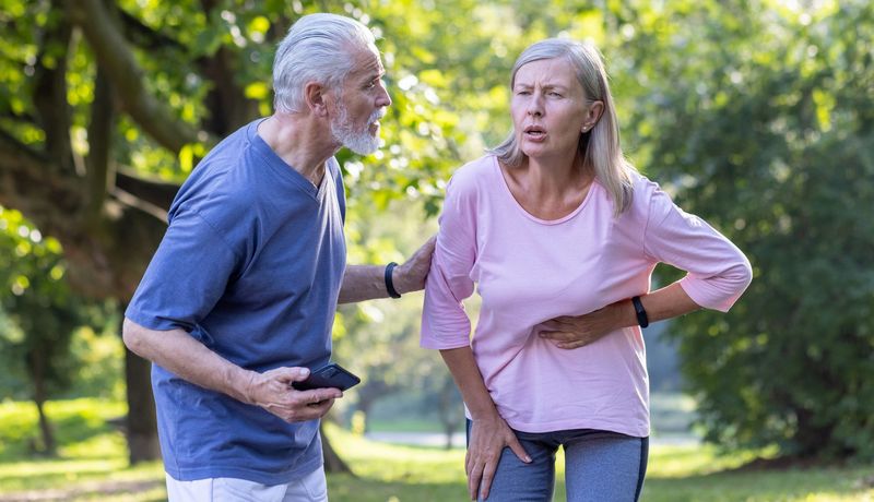 Ältere Frau hält sich den Oberbauch, ein Mann steht erschrocken neben ihr, das Handy in der Hand.