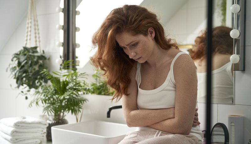 Junge Frau blickt sorgenvoll in Unterhemd und Schlafanzughose im Badezimmer, die Arme vor dem Bauch verschränkt. 