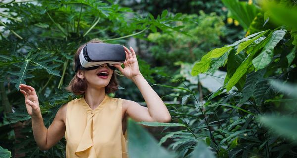Eine junge Frau trägt mit freudigem Gesichtsausdruck eine VR-Brille inmitten tropischer Pflanzen.