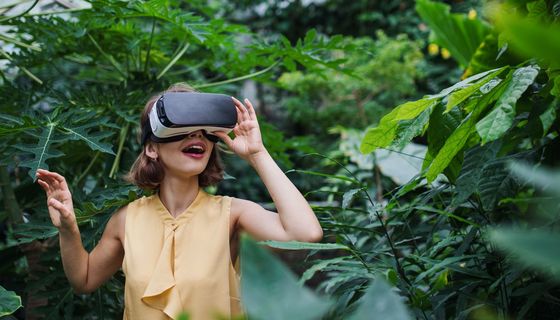 Eine junge Frau trägt mit freudigem Gesichtsausdruck eine VR-Brille inmitten tropischer Pflanzen.