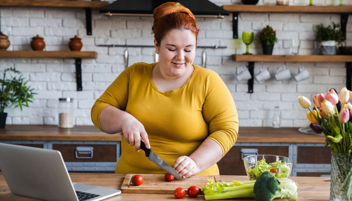 Eine junge, übergewichtige Frau steht in der Küche, schneidet Tomaten und macht sich einen Salat.
