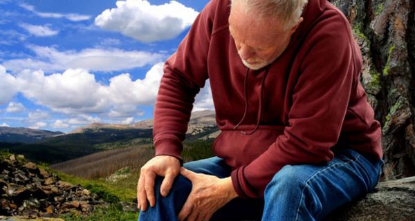 Senior in den Bergen auf Fels sitzend und sich ans rechte Knie greifend