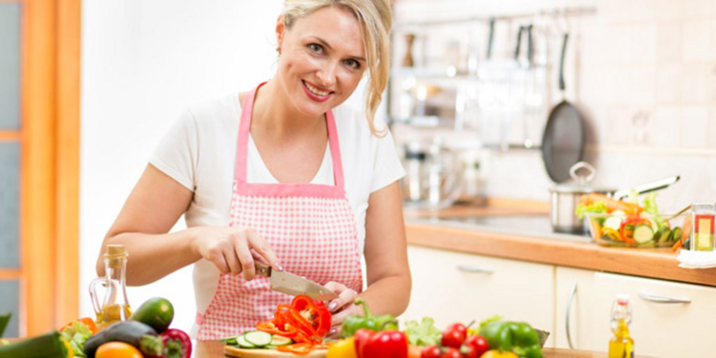 Blonde Frau um die 40 mit rotweißkarierter Schürze beim Gemüseschneiden in der Küche. Sie lächelt in die Kamera